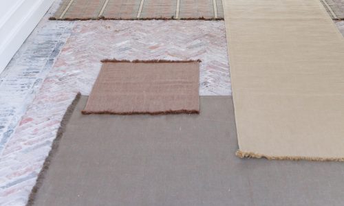 Mehrere rechteckige Teppiche in gedämpften Braun-, Hellbraun- und Grautönen sind auf einem gemusterten Ziegelsteinboden ausgelegt, wobei sich einige Teppiche überlappen. Im Hintergrund ist ein Teil einer weißen Wand zu sehen.