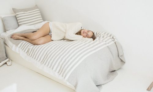 Eine Frau in einem cremefarbenen Pullover und Shorts entspannt sich auf einem gestreiften Bett in einem minimalistischen weißen Zimmer, das mit dem Camille Leinen Plaid von Libeco drapiert ist, und blickt nachdenklich in die Ferne.