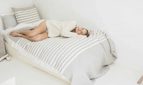 Eine Frau in einem cremefarbenen Pullover und Shorts entspannt sich auf einem gestreiften Bett in einem minimalistischen weißen Zimmer, das mit dem Camille Leinen Plaid von Libeco drapiert ist, und blickt nachdenklich in die Ferne.