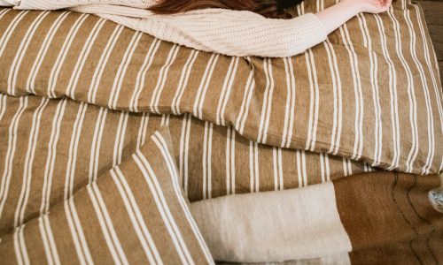 Eine Frau in einem cremefarbenen Pullover liegt entspannt auf einem Bett mit braunem und weißem Canal Stripe Leinen Bettwäsche von Libeco, die Arme über den Kopf gestreckt.