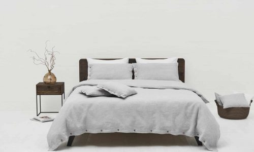 A neatly made bed with Caroline Buttons Linen Bedding by Geniksa in light grey and two pillows stands against a white wall. To the left is a dark wooden nightstand with a vase of dried branches, to the right a basket and a folded blanket.