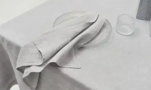 Ein Teller und ein Becher aus Klarglas stehen auf einer hellgrauen Tischdecke, über der eine passende graue Leinenserviette drapiert ist.