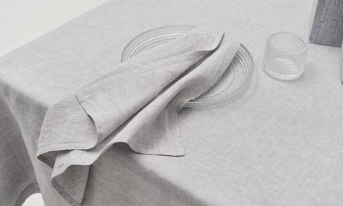 On the Caroline linen tablecloth by Geniksa, a clear glass plate holds a folded light grey napkin, accompanied by a small textured glass tumbler for a clean, minimalist table setting.