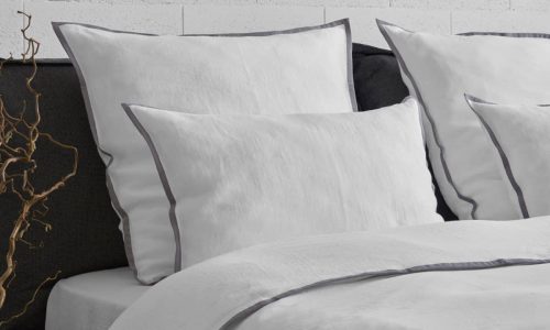 Ein ordentlich gemachtes Bett mit weißem Bettzeug und grauen Paspeln an den Rändern, vor einer hellen, strukturierten Wand. Neben dem Bett sind ein dunkelgraues Kopfteil und eine große Glasvase mit getrockneten Zweigen zu sehen.