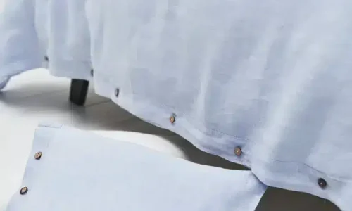 Close-up of a light blue Cygna Classic Linen Sheet by Geniksa with brown edge buttons, shown with a matching pillow and bedspread on a white floor.