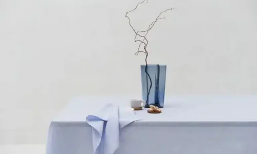 Ein minimalistisch gedeckter Tisch mit einer hellblauen Tischdecke, einer blauen Glasvase mit kahlen Zweigen, einer kleinen weißen Tasse und ein paar verstreuten Keksen, alles vor einem schlichten weißen Hintergrund.