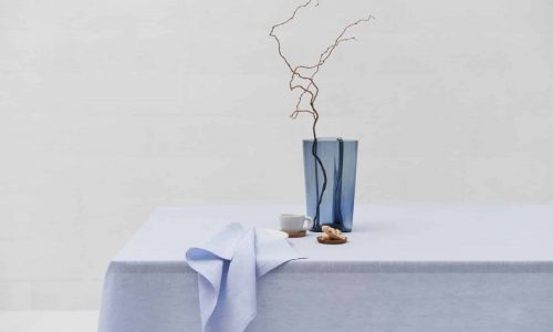A minimalist table setting with the Celeste linen tablecloth by Geniksa, a blue glass vase with a twisted branch, a folded napkin, a small white bowl, a white cup and biscuits on a neutral background.