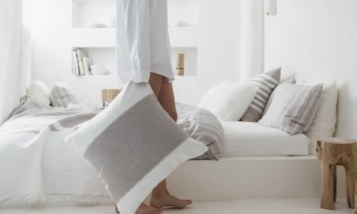 Eine Person in einem weißen Hemd hält ein grau-weißes Charlotte Leinen Plaid von Libeco-Kissen und steht barfuß neben einem ordentlich gemachten Bett in einem hellen, minimalistischen Schlafzimmer mit neutralen Tönen und sanfter Beleuchtung.