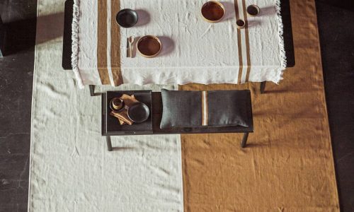 Draufsicht auf einen rechteckigen Esstisch mit weißer Tischdecke und hellbraunen Streifen, bestückt mit Keramikgeschirr und Tassen. Die Bank und der Teppich unter dem Tisch passen zu dem weiß-hellbraunen Farbblockmuster der Tischdecke.