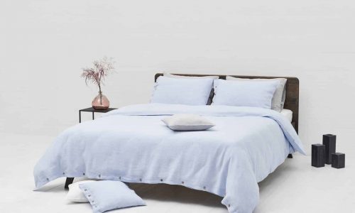 A neatly made bed with light blue Cygna Buttons Linen Bedding by Geniksa stands in a minimalist room, beside a small table with a pink vase of dried flowers, with two black decorative objects on the floor nearby.