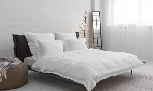 Minimalist bedroom with a low bed with white bedding and pillows, a brown stool with a blanket, a floor mirror, a bare decorative tree with lights and a large window with sheer curtains.