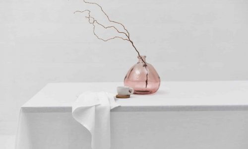 A minimalist scene with the Cygna linen tablecloth by Geniksa, a pink glass vase with bare branches, a white cup and saucer and a draped napkin, all against a plain white background.