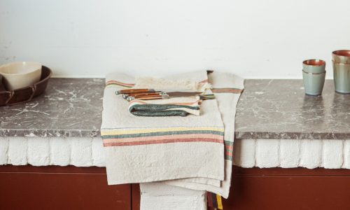 Ein gefaltetes gestreiftes Tuch mit Messern und einem Gemüseschäler liegt auf einer Marmorarbeitsplatte. Zwei Keramiktassen und eine Schüssel stehen daneben an einer schlichten weißen Wand.