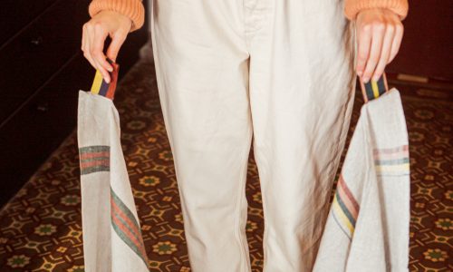 Eine Person in einem pfirsichfarbenen Pullover und einer cremefarbenen Hose hält zwei bunt gestreifte, wiederverwendbare Taschen - eine mit dem Design Dock Green Stripe - reines Leinen für die Küche von Libeco. Darunter ist ein gemusterter Fliesenboden zu sehen.