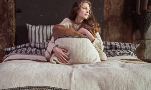 Eine Frau mit langen braunen Haaren sitzt auf einem Bett, das mit erdfarbenen und gestreiften Decken bedeckt ist, darunter die Etienne Leinen Coverlet und Kissen von Libeco. Sie schmiegt sich an ein großes Kissen und blickt nachdenklich zur Seite in einem sanft beleuchteten, rustikalen Raum.