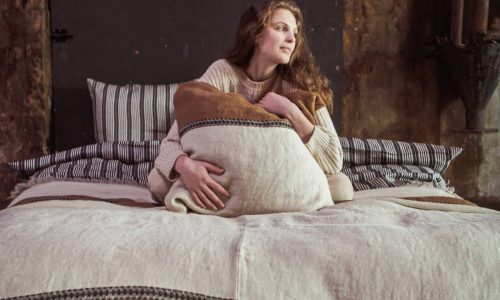 Eine Frau mit langen braunen Haaren sitzt auf einem Bett, das mit erdfarbenen und gestreiften Decken bedeckt ist, darunter die Etienne Leinen Coverlet und Kissen von Libeco. Sie schmiegt sich an ein großes Kissen und blickt nachdenklich zur Seite in einem sanft beleuchteten, rustikalen Raum.