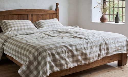 Ein rustikales Schlafzimmer mit einem hölzernen Bettgestell und passender karierter Bettwäsche, die auf einem Holzboden neben einem Fenster mit Eisenstangen steht. Eine braune Vase mit getrockneten Gräsern steht auf der Fensterbank.