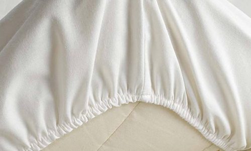 Eine Hand spannt den elastischen Rand eines weißen Spannbetttuchs über die Ecke einer Matratze. Die Oberfläche darunter ist eine gewebte Matte.