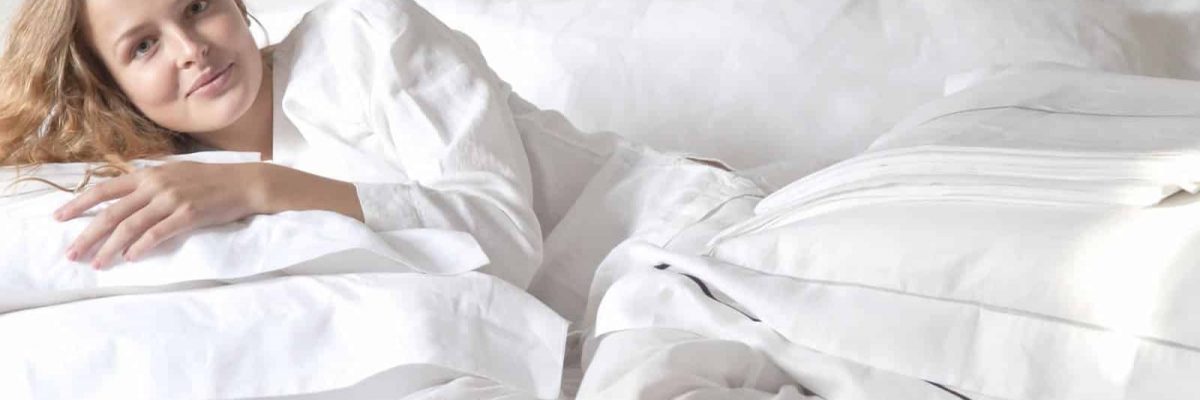 Eine Frau mit hellem Haar und weißem Schlafanzug liegt auf einem Bett mit weißen Laken und Kissen und lächelt sanft in die Kamera in einem hellen, minimalistischen Raum.