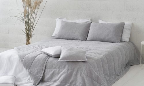 Ein ordentlich gemachtes Bett mit hellgrauer Bettwäsche und Kissen präsentiert die Caroline Leinen Tagesdecke von Geniksa vor einer weiß strukturierten Wand. Eine Vase mit hohem getrocknetem Gras verleiht dem Schlafzimmer Ruhe und einen minimalistischen Stil.