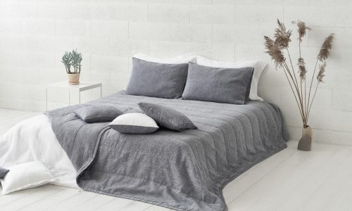 Ein minimalistisches Zimmer mit einem ordentlich gemachten Bett mit grauer Heron Classic Leinen Tagesdecke von Geniksa, weißen Akzenten, einer weißen Backsteinwand, einem kleinen Beistelltisch mit einer Topfpflanze und hohen getrockneten Gräsern in einer Vase.