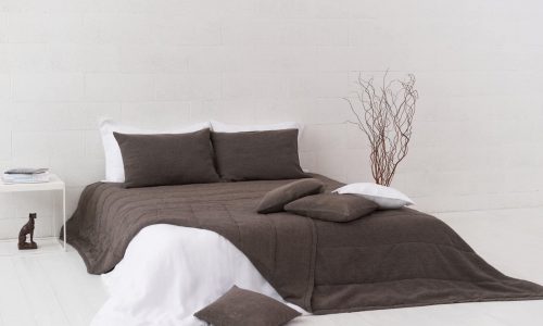 Ein ordentlich gemachtes Bett mit dunkelgrauer Bettwäsche, darunter die Jungle Classic Leinen Tagesdecke von Geniksa, weiße Laken, drei passende Kissen auf dem Bett und zwei auf dem Boden. Auf einem weißen Beistelltisch stehen eine kleine Skulptur und Bücher in einem minimalistischen weißen Raum.