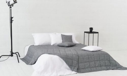 Ein minimalistisches Schlafzimmer mit einem niedrigen Bett in Grau und Weiß, mit der Scotia Leinen Tagesdecke von Geniksa, drei Kissen, einem schwarzen Beistelltisch mit einem kleinen Gegenstand und einer Studiolampe auf einem Ständer vor weißen Wänden und Böden.