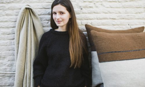 Eine junge Frau mit langen braunen Haaren in einem schwarzen Pullover steht vor einer weißen Backsteinwand, neben einer beigen Hängedecke und zwei großen braunen und beigen Kissen aus Gus Leinen-Wolle-Mix von Libeco.