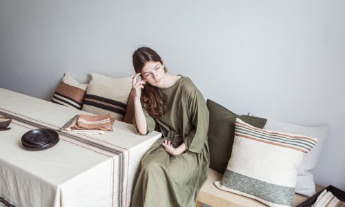 Eine Frau in einem olivgrünen Kleid sitzt auf einer Holzbank neben einem Tisch mit gestreifter Bettwäsche und Kissen aus Gips - reines Leinen für den Tisch von Libeco, stützt ihren Kopf auf die Hand und blickt nachdenklich nach vorn.