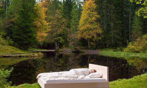A person sleeps outdoors beside a tranquil forest pond on a bed with the Pure Bamboo Summer Duvet by HEFEL, surrounded by lush greenery and autumn-coloured trees.