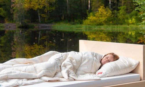 Eine Person schläft friedlich auf einer Sommerdecke aus reiner Wolle von HEFEL im Freien an einem ruhigen See, eingehüllt in weiße Bettwäsche und umgeben von grünen und gelben Bäumen.