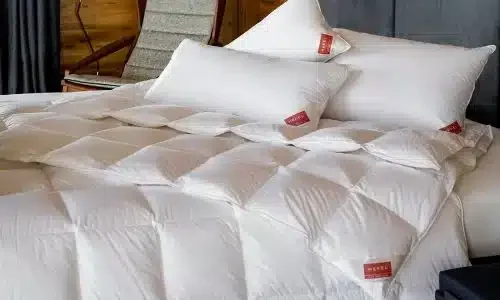 Ein ordentlich gemachtes Bett mit der KlimaControl Comfort Winterdecke von HEFEL - weißes, gestepptes Bettzeug mit zwei großen weißen Kissen, die jeweils mit einem roten Etikett versehen sind. Ein dunkles Kopfteil, ein Sessel und eine Holzvertäfelung sorgen für einen eleganten Hintergrund.
