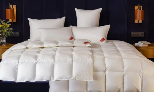 A neatly made bed mit einer whiten, quiltedn duvet und fünf whiten pillow, von denen zwei rote Etiketten haben. Das Bett steht vor einem dunklen, upholstered headboard mit braunen Wandlampen auf jeder Seite.