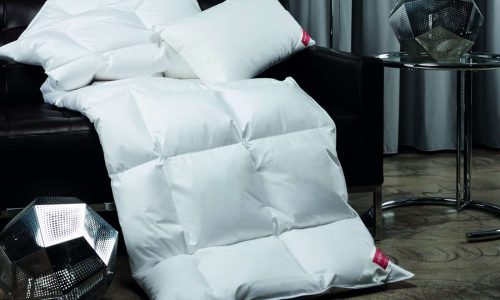 Weiße Kissen und eine bauschige weiße Bettdecke liegen auf einem dunklen Sofa und sind auf dem Holzboden drapiert; daneben stehen zwei geometrische Metalldekorationen und ein runder Beistelltisch aus Glas, mit grauen Vorhängen im Hintergrund.