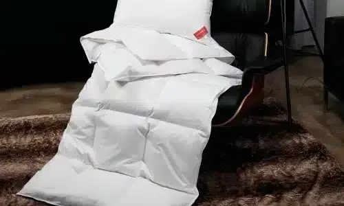 Eine KlimaControl Comfort Winterdecke von HEFEL und ein Kissen liegen auf einem schwarzen Ledersessel, darunter ein brauner Kunstfellteppich, der eine gemütliche und einladende Atmosphäre schafft.