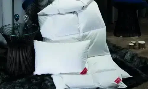 Ein weißes Kissen und die KlimaControl Comfort Winterdecke von HEFEL liegen auf einem dunklen Kunstfellteppich neben einem modernen Glasbeistelltisch, im Hintergrund sind Sanduhren und Stühle zu sehen.