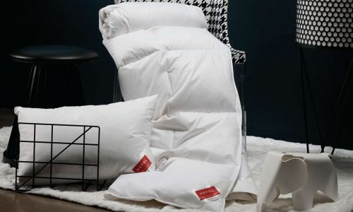 Eine gemütliche Szene mit der Daune DeLuxe Daunen-Ganzjahresdecke von HEFEL, einer weißen Bettdecke, einem Kissen und einem Kissen mit roten Anhängern auf einem flauschigen Teppich sowie einem schwarzen Hocker, einem Stuhl mit Hahnentrittmuster, einem Drahtkorb und einem Dekor in Elefantenform.