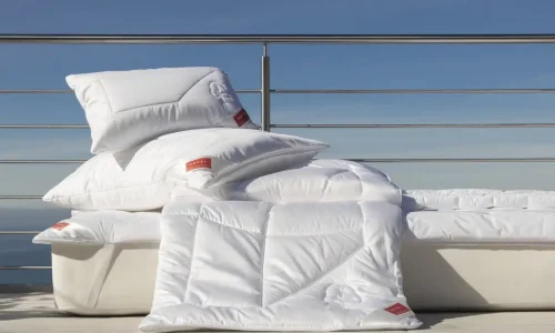 A stack of white pillows and a white duvet from the renowned linen weaving mills, all bearing red labels, neatly placed on a white outdoor lounge chair against a backdrop of clear blue sky and metal railings.