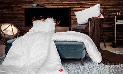 Ein gemütliches Wohnzimmer mit einem modernen Kamin, einem tealfarbenen Ottomanen, der mit der KlimaControl Comfort Winterdecke von HEFEL drapiert ist, einem Kissen auf einem braunen Sessel, einer braunen Decke, einem geometrischen blau-weißen Teppich und einer warmen Umgebungsbeleuchtung zum Entspannen.