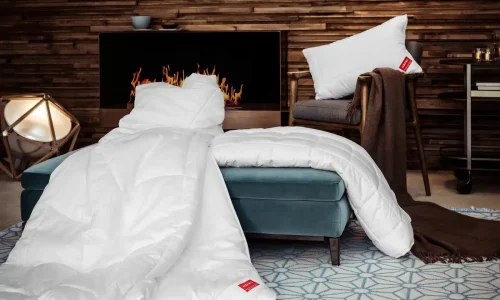 Ein gemütliches Wohnzimmer mit einem modernen Kamin, einem blauen Hocker mit der KlimaControl Comfort Winterdecke von HEFEL, einem weißen Kissen auf einem braunen Stuhl und einer weichen Decke auf einem geometrisch gemusterten Teppich.