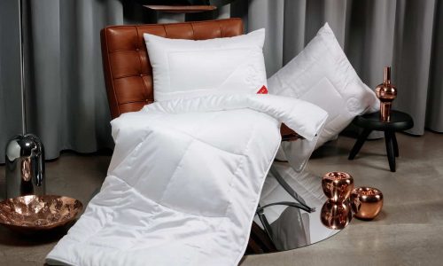 On a brown leather armchair lie a white pillow and a duvet, another white pillow lies on a black stool next to copper decorative items and a grey curtain in the background.