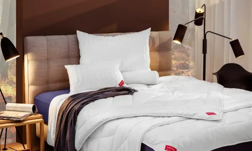 A neatly made bed with an upholstered headboard, white bedding and pillows, a dark throw, two modern black floor lamps and a window with sheer curtains letting in natural light.