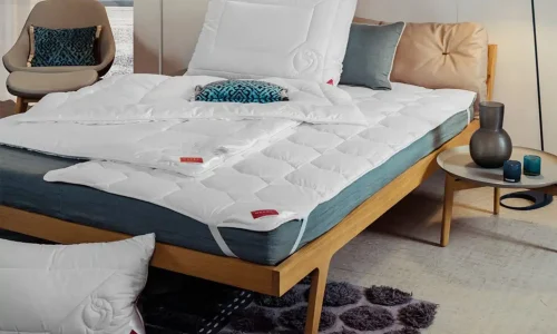 A modern bedroom with a wooden bed frame, a blue mattress, white quilted bedding, pillows, a beige headboard, an accent chair, decorative cushions, a side table with vases and a textured rug on the floor.