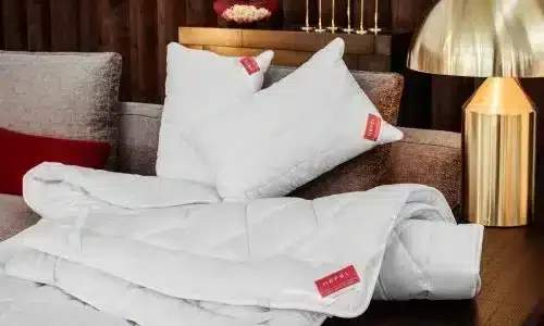 Weiße Kissen und eine gefaltete KlimaControl Comfort Winterdecke von HEFEL mit rotem Etikett sind auf einem beigen Sofa angeordnet, während eine goldene Lampe und dekorative Akzente im warm beleuchteten Hintergrund erscheinen.