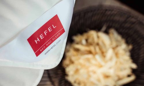 Eine Nahaufnahme einer weißen Stoffecke mit rotem "Hefel Bed and Sleep"-Etikett über einem unscharfen Korb aus leicht zerkleinertem Material zeigt die Bio Zirbe Ganzjahresdecke von HEFEL.