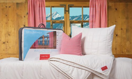 Ein gemütliches Schlafzimmer mit holzgetäfelten Wänden, roten Gingham-Vorhängen und einem Fenster, das verschneite Berge zeigt. Auf dem Bett liegen eine weiße Bettdecke, ein Kissen und ein dazu passendes kariertes Kissen. Die verpackte Bettwäsche ist mit HEFEL beschriftet.