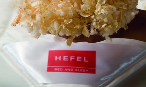 Eine Hand hält eine Mischung aus weichem weißem und hellbraunem Füllmaterial über einer weißen Bettdecke mit einem roten Etikett mit der Aufschrift HEFEL BED AND SLEEP.