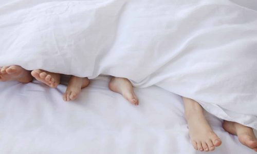 Vier Fußpaare lugen unter einer weißen Decke auf einem Bett hervor, was darauf hindeutet, dass mehrere Personen zusammenliegen und sich entspannen. Das weiße Bettzeug schafft eine weiche und gemütliche Atmosphäre.