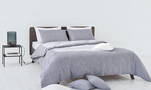 In a minimalist room, a neatly made bed with Heron Classic Linen Bedding by Geniksa in grey is complemented by white pillows. Next to the bed is a nightstand with a lamp and a book, and pillows are scattered on the white wooden floor.