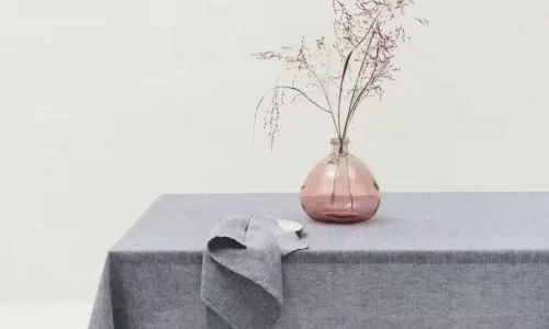 Eine einfache Szene mit einem grauen Tischtuch, das einen Tisch bedeckt, einer grauen Serviette, die über die Seite drapiert ist, und einer rosafarbenen Glasvase mit spärlichen getrockneten Zweigen, alles vor einem schlichten weißen Hintergrund.